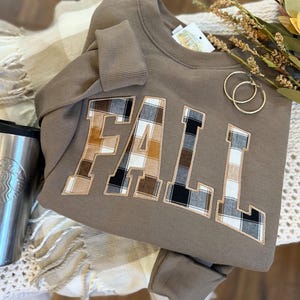 May include: A mocha brown crewneck sweatshirt featuring the word FALL in large block letters. Each letter is filled with a brown, black, and white plaid fabric pattern and outlined with a tan border. The sweatshirt has ribbed cuffs and a collar.