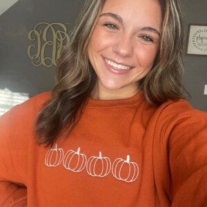 May include: A woman wearing a burnt orange sweatshirt with a white embroidered design of four pumpkins.