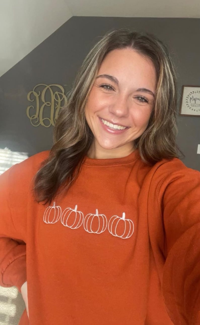 May include: A woman wearing a burnt orange sweatshirt with a white embroidered design of four pumpkins.