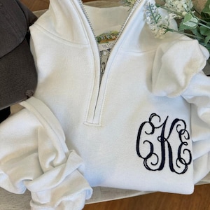 May include: White quarter-zip sweatshirt with a silver zipper and a navy blue monogram. The sweatshirt has long sleeves and a high collar. A dark gray baseball cap is visible in the background.