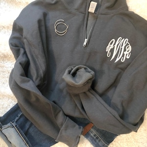 May include: A charcoal gray quarter-zip sweatshirt with a white monogram on the front. The sweatshirt has a silver zipper and rolled-up sleeves. A pair of silver hoop earrings and blue jeans are also in the image.