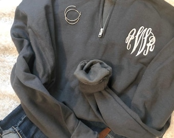 May include: A charcoal gray quarter-zip sweatshirt with a white monogram on the front. The sweatshirt has a silver zipper and rolled-up sleeves. A pair of silver hoop earrings and blue jeans are also in the image.