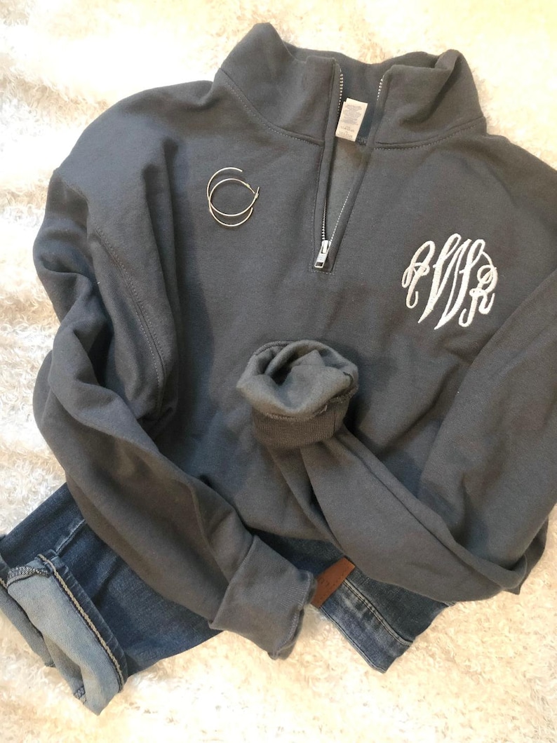 May include: A charcoal gray quarter-zip sweatshirt with a white monogram on the front. The sweatshirt has a silver zipper and rolled-up sleeves. A pair of silver hoop earrings and blue jeans are also in the image.