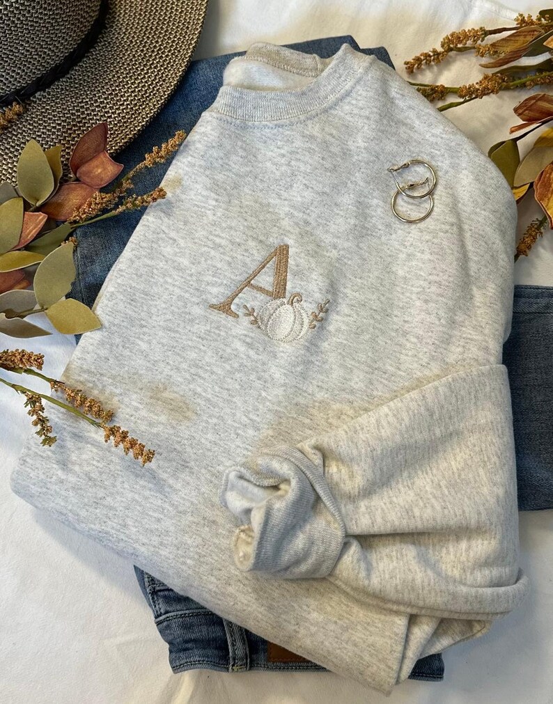 May include: A grey sweatshirt with a brown embroidered letter A and a white pumpkin design. The sweatshirt is folded and displayed on a white surface.