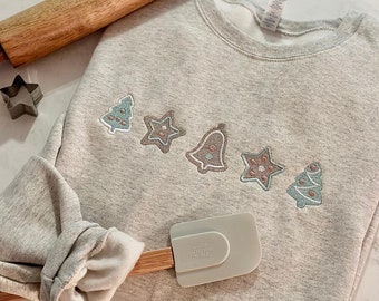 Può includere: Una felpa grigia con un designo festivo di biscotti di pan di zenzero, tra cui una stella, una campana e un albero. La felpa è appoggiata su una superficie bianca con un mattarello di legno e una spatola.