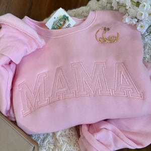 May include: A light pink sweatshirt with the word "MAMA" embroidered across the front in a block letter style. Gold hoop earrings with heart details rest on the sweatshirt. A tag with a house illustration is visible.