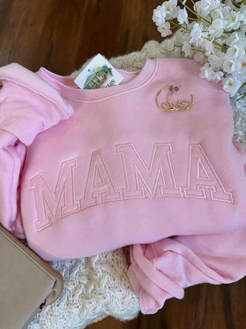 May include: A light pink sweatshirt with the word "MAMA" embroidered across the front in a block letter style. Gold hoop earrings with heart details rest on the sweatshirt. A tag with a house illustration is visible.