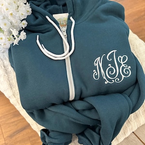 May include: Teal zip-up hoodie with white drawstring and a silver zipper. The hoodie features a white embroidered monogram on the front. The hoodie is folded on a white textured surface, with white flowers in the upper left corner.