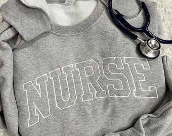 May include: Gray sweatshirt with the word "NURSE" in white outlined letters. A blue stethoscope is lying on top of the sweatshirt.