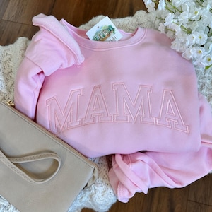 May include: A light pink sweatshirt with the embroidered word "MAMA" in a matching pink thread. The sweatshirt is folded on top of a cream-colored knitted blanket, with a light beige clutch and white flowers in the background.