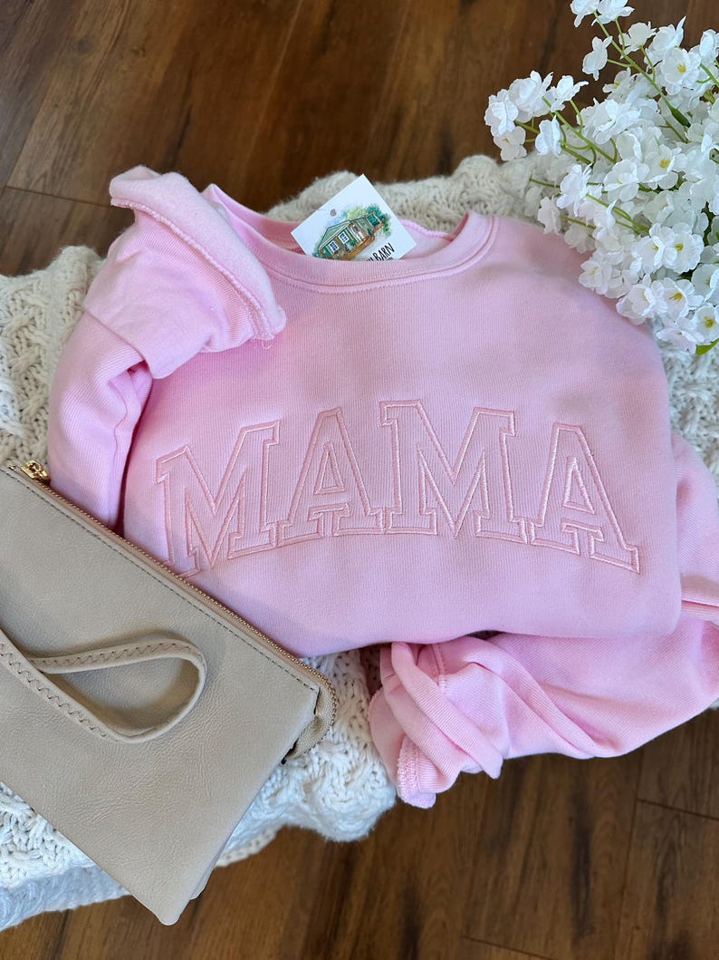 May include: A light pink sweatshirt with the embroidered word "MAMA" in a matching pink thread. The sweatshirt is folded on top of a cream-colored knitted blanket, with a light beige clutch and white flowers in the background.