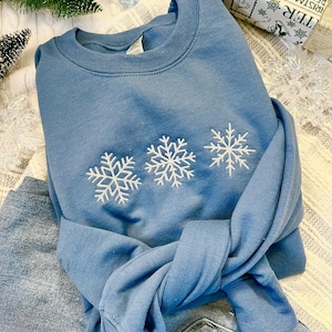 May include: A blue sweatshirt with three white embroidered snowflakes on the front. The sweatshirt is folded and lying on a pair of blue jeans.