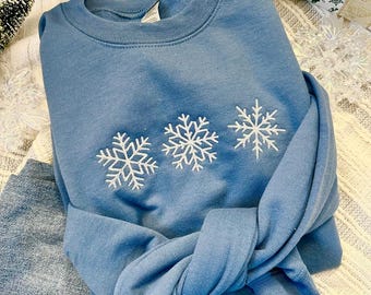 Può includere: Una felpa blu con tre fiocchi di neve ricamati in bianco sul davanti. La felpa è piegata e appoggiata su un paio di jeans blu.