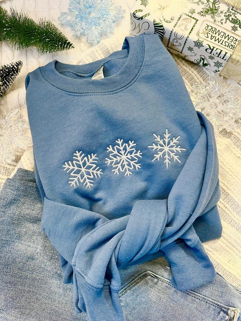 May include: A blue sweatshirt with three white embroidered snowflakes on the front. The sweatshirt is folded and lying on a pair of blue jeans.