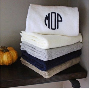May include: A stack of towels in various colors, including white, gray, beige, and navy blue. The top towel has a white background with a black circle containing the monogram "MOP".