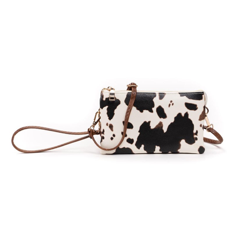 Puede incluir: Un peque&ntilde;o bolso bandolera blanco y negro con estampado de vaca con una correa marr&oacute;n. El bolso tiene una cremallera y un tirador de cremallera dorado.
