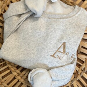 May include: A grey sweatshirt with a brown embroidered letter "A" and a pumpkin design. The sweatshirt is folded and displayed on a woven basket.
