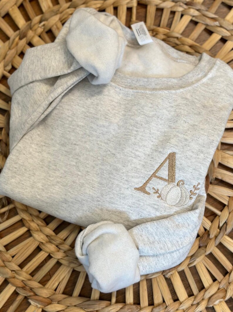 May include: A grey sweatshirt with a brown embroidered letter "A" and a pumpkin design. The sweatshirt is folded and displayed on a woven basket.