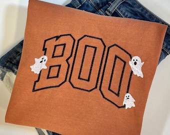 May include: A burnt orange sweatshirt with the word "BOO" embroidered in black outlined letters. Three white ghost appliques are placed around the letters.