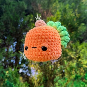 May include: A small, round, orange crocheted plush toy with a textured surface. It features two black plastic eyes, a simple horizontal stitched mouth, a small tan stem on top, and a ruffled light green leaf accent on the side.