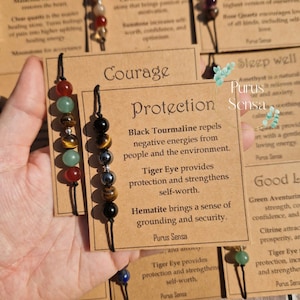 May include: A brown card with the text "Courage Protection" and a description of the benefits of black tourmaline, tiger eye, and hematite. The card is next to a bracelet with black, green, brown, and red beads.