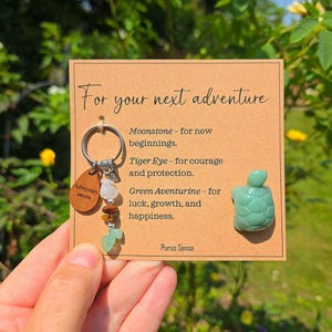 May include: A keyring with a wooden tag reading "Adventure awaits" and various gemstones. The card says "For your next adventure" and lists the properties of the stones. A small green turtle figurine is also present.