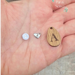 May include: Three small charms with the letter "A" displayed on a hand. The charms include a white bead, a silver heart, and a wooden teardrop shape. The wooden charm has a dark brown engraved letter.
