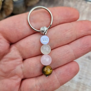 May include: A silver keychain with a charm of four gemstone beads in light blue, white, pink, and brown. The top bead has the letter "W". The keychain is held in a hand.