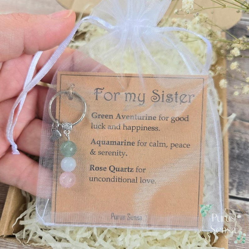 Pode incluir: Um chaveiro com um anel prateado e tr&ecirc;s contas de pedras preciosas: aventurina verde, &aacute;gua-marinha branca e quartzo rosa. O chaveiro est&aacute; em um saco de organza branco com uma etiqueta que diz "Para minha irm&atilde;".