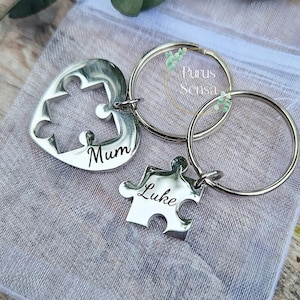 Op de afbeelding: Twee zilveren puzzelstukjes sleutelhangers met de namen "Mum" en "Luke" erop gegraveerd. De sleutelhangers zijn bevestigd aan zilveren sleutelringen.