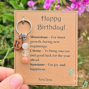 May include: A keychain with a silver ring, wooden letter "R" charm, and three gemstone beads. The card reads "Happy Birthday!" with descriptions of the gemstones: Moonstone, Citrine, and Sunstone. The brand name "Purus Sensa" is also visible.