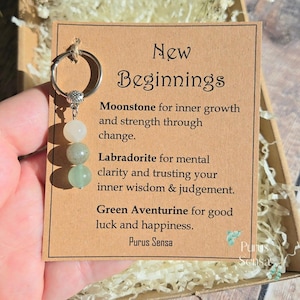 Può includere: Un portachiavi argentato con tre perline di pietre preziose: pietra di luna bianca, labradorite grigia e avventurina verde. Il portachiavi è attaccato a una carta con il testo "New Beginnings" e descrizioni delle pietre preziose.