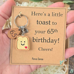 Pode incluir: Um porta-chaves com uma etiqueta de madeira com o número 65, um pingente de torrada com um rosto sorridente e um coração, e um cartão que diz "Here's a little toast to your 65th birthday! Cheers!"