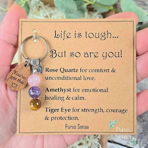 Peut inclure: Un porte-clés avec une étiquette en bois disant "You've got this" et un cœur. Il comporte un anneau argenté et trois perles de pierres précieuses : quartz rose, améthyste et œil de tigre. La carte dit "Life is tough... But so are you!"