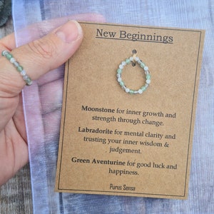 Könnte beinhalten: Eine braune Karte mit dem Text "Neuanfänge" und Beschreibungen der Steine in einem Armband: Mondstein für inneres Wachstum und Stärke durch Veränderung, Labradorit für mentale Klarheit und Vertrauen in die eigene innere Weisheit und das eigene Urteilsvermögen, grüner Aventurin für Glück und Zufriedenheit. Die Karte enthält auch den Text "Purus Sensa".