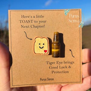 Può includere: Una carta marrone con il testo "Here's a little TOAST to your Next Chapter!" e "Tiger Eye brings Good Luck & Protection." Presenta un ciondolo a forma di toast con una faccina sorridente e un cuore, e una pietra occhio di tigre.