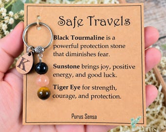 May include: A metal keychain featuring a wooden charm with the letter K and three round beads in black, peach, and brown. The keychain is attached to a tan card with the text Safe Travels, Black Tourmaline, Sunstone, Tiger Eye, and Purus Sensa.