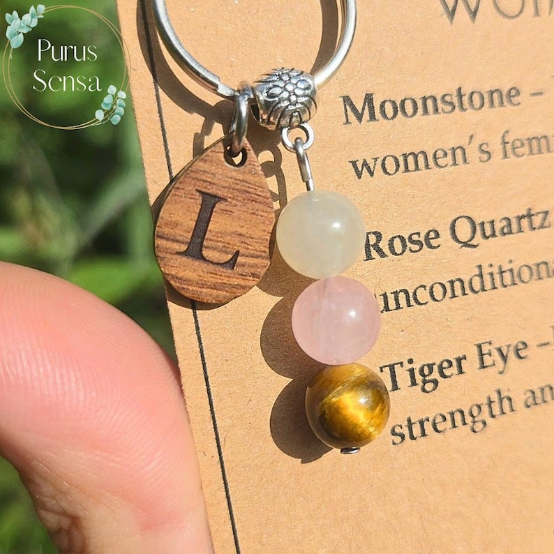 May include: A silver keyring with a wooden teardrop pendant engraved with the letter "L". The pendant is attached to a charm with three gemstone beads: a light green, a pink, and a gold-brown tiger eye bead. The text "Purus Sensa" is in the upper left corner.