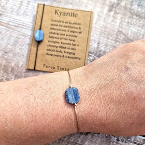 May include: A blue kyanite stone bracelet on a brown string. The bracelet is on a person's wrist. The card reads "Kyanite is an excellent stone for meditation & attunement, it aligns all chakras and provides balance of Yin-Yang energies. Kyanite has a calming effect on the whole body, bringing deep peace & tranquility. Purus Sensa"