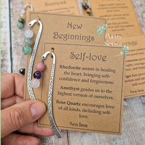 May include: A silver bookmark with a decorative design and a chain of pink, purple, and green gemstones. The bookmark is attached to a card with the text "New Beginnings Self-love" and a description of the stones' properties. The card is brown with a white border.