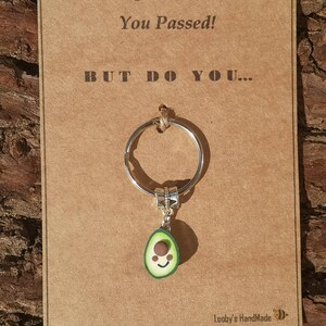 May include: A brown card with the text "Congratulations You Passed! BUT DO YOU..." and a silver key chain with a green avocado charm. The avocado charm has a brown seed and a smiling face.