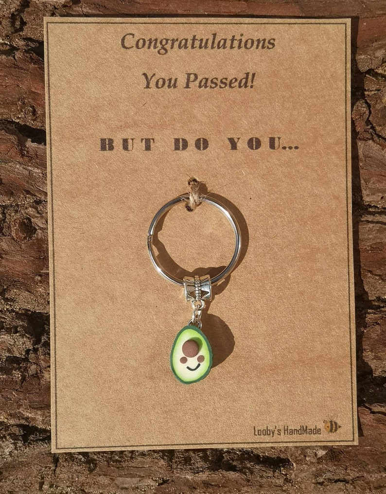 May include: A brown card with the text "Congratulations You Passed! BUT DO YOU..." and a silver key chain with a green avocado charm. The avocado charm has a brown seed and a smiling face.