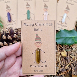 Può includere: Un biglietto marrone con il testo "Merry Christmas Katie" e un folletto dei cartoni animati che tiene un cristallo di occhio di tigre. Il testo "Protection Elf" e "Purus Sensa" è anche sulla carta.