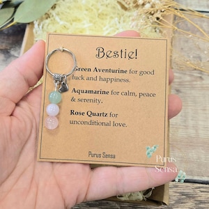May include: A silver-toned metal keyring featuring three round gemstone beads in green, pale blue, and light pink, attached to a card with the text: Bestie! Green Aventurine for good luck and happiness. Aquamarine for calm, peace & serenity. Rose Quartz for unconditional love. Purus Sensa.