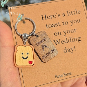 May include: A keychain with a toast-shaped charm featuring a smiling face and a red heart. A wooden tag with "Cheers!" and the date 01.09.26 hangs next to it. The card reads "Here's a little toast to you on your Wedding day!"