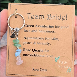 May include: A brown card with the text "Team Bride!" and a description of the meaning of each stone on a keychain. The stones are green aventurine, aquamarine, and rose quartz. The text on the card reads: "Green Aventurine for good luck and happiness. Aquamarine for calm, peace & serenity. Rose Quartz for unconditional love. Purus Sensa"