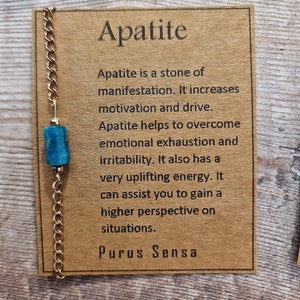 May include: A gold chain necklace with a blue apatite stone pendant. The card reads "Apatite is a stone of manifestation. It increases motivation and drive. Apatite helps to overcome emotional exhaustion and irritability. It also has a very uplifting energy. It can assist you to gain a higher perspective on situations. Purus Sensa"