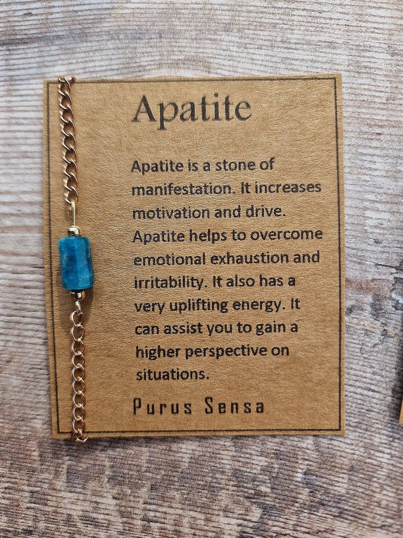 May include: A gold chain necklace with a blue apatite stone pendant. The card reads "Apatite is a stone of manifestation. It increases motivation and drive. Apatite helps to overcome emotional exhaustion and irritability. It also has a very uplifting energy. It can assist you to gain a higher perspective on situations. Purus Sensa"