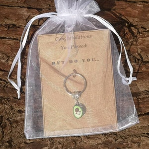May include: A white organza bag with a brown card that reads "Congratulations You Passed! But Do You..." and a silver avocado keychain with a green avocado charm.