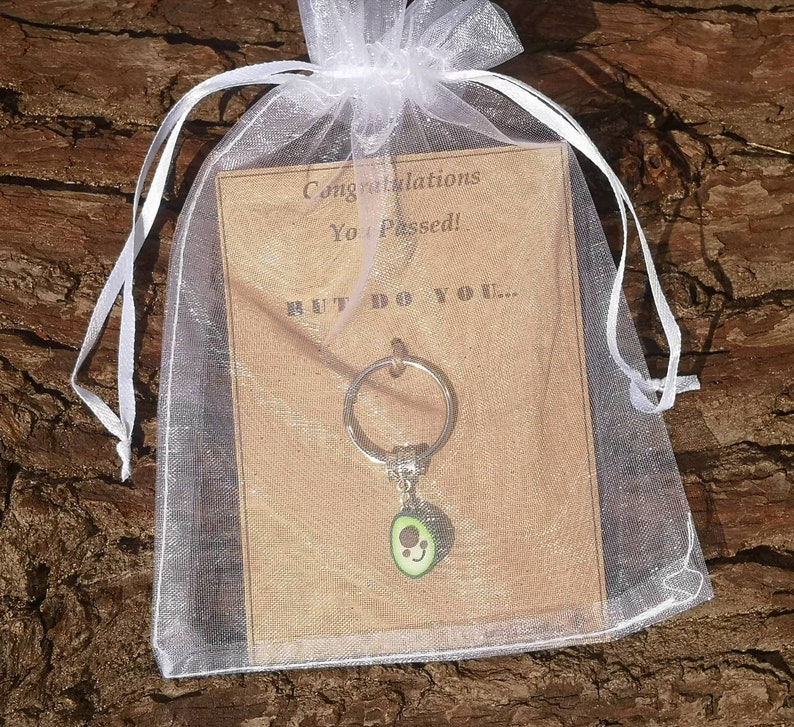 May include: A white organza bag with a brown card that reads "Congratulations You Passed! But Do You..." and a silver avocado keychain with a green avocado charm.